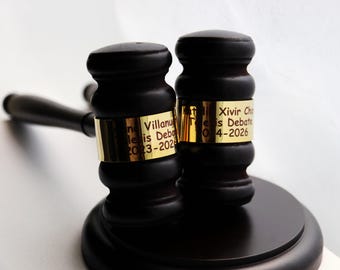 May include: A black gavel with a round base and a handle. The gavel head has a gold band with engraved text. The text includes names and dates, likely for a debate or legal event. The gavel is set against a white background.
