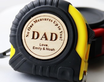 May include: A yellow and black retractable tape measure featuring a circular wooden inlay on the side. The wood is engraved with the text No One Measures Up to You! DAD Love, Emily & Noah in a dark brown font, centered on the tool casing.