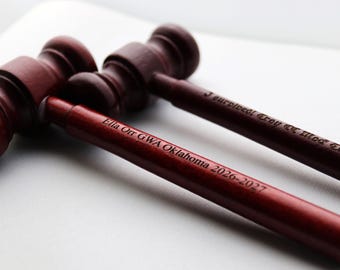 May include: Three wooden gavels with dark red stain. The gavel handles are engraved with text. One reads "Ella Orr GWA Oklahoma 2026-2027" and another reads "J europecd Troy et Mod Trial 20". The gavels are on a white surface.