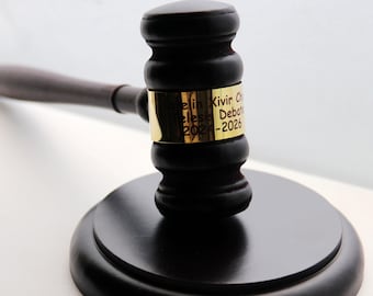 May include: A dark brown wooden gavel with a polished finish and a gold-colored metal band wrapped around the head. The band features black engraved text that reads "Kaelin Xivir Chair" followed by "Telesis Debate" and "2024-2026" on three lines.