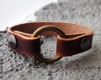 Puede incluir: Un brazalete de cuero marrón con un anillo de latón y dos detalles metálicos.