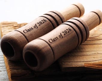 May include: Two wooden noisemakers with the text "Class of 2026" engraved on them. The noisemakers are cylindrical with grooves and rounded ends. They rest on a wooden surface, suggesting a graduation theme.