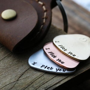 May include: Three guitar picks in silver, copper, and gold, each stamped with "I pick you." A brown leather case with a brass button is in the background. The picks rest on a wooden surface.