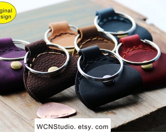 May include: A set of seven leather guitar pick holders in various colors, including purple, brown, tan, black, and red. Each holder has a silver ring and a brass button. The holders are arranged on a wooden surface with a copper guitar pick that says "I pick you" in the center.