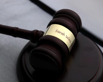 May include: A dark brown wooden gavel with a matching base. A gold band on the gavel reads "Sarah Wins." The gavel is resting on its base, suggesting a moment of finality or celebration.