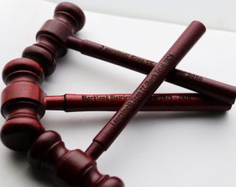 May include: Four dark red wooden gavels are arranged on a white surface. The gavels have engraved text on the handles, including "Blackland District" and "Surrinnell Troy CC Mock Trial 2026". The gavels are a deep reddish-brown color.