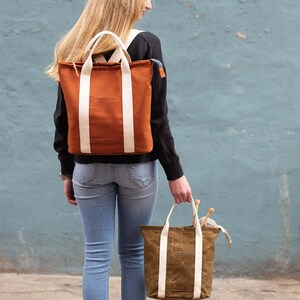 Może przedstawiać: Osoba pokazana od tyłu, nosi plecak w kolorze rdzy i pasującą torbę. Obie torby mają kremowe paski, a u góry znajduje się napis "BUCKTHORN backpack & tote". Torba jest wypełniona akcesoriami do robienia na drutach.