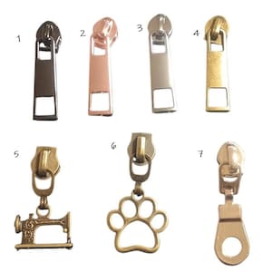 May include: A collection of seven metal zipper pulls labeled 1 through 7. Pulls 1 through 4 are rectangular with cutouts. Pull 5 is a sewing machine charm, pull 6 is a paw print charm, and pull 7 is a rounded rectangular pull with a circular cutout.