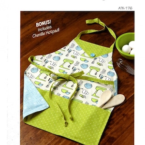 May include: An apron with a green and white pattern featuring kitchen utensils and appliances. The apron has a green trim and ties, with a blue button detail. The text "MOM AND ME APRONS" is visible.