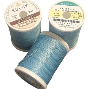 May include: Three spools of light blue cotton thread. Two spools stand upright, while one lies horizontally in front. The labels read Sulky Cotton 30, noting long staple, matte finish, colorfast, 30 wt., needle 90, and 500 yds. The thread is a solid light blue color.