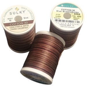 May include: Three spools of Sulky Cotton 30 Blendables thread in a variegated brown color. Each spool features white plastic ends with printed text including brand name, 30 wt., 500 yards, and 100% cotton. The thread has a matte finish.