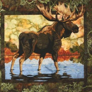 May include: A framed fabric art piece featuring a moose standing in water. The moose has large antlers and is depicted in shades of brown and black. The background shows a landscape with water and trees, framed by a border of green foliage.
