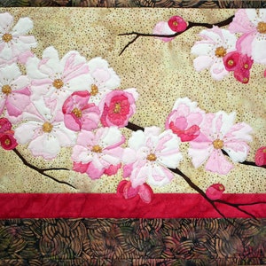 May include: A fabric art piece featuring a floral design. The artwork showcases white and pink blossoms with yellow centers, set against a beige background. Dark brown branches and a red border frame the design, with a patterned edge.