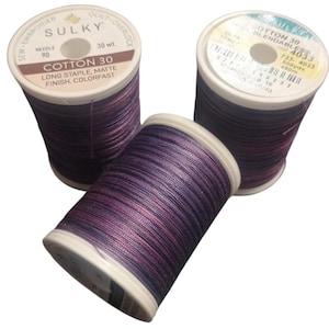 May include: Three spools of Sulky Cotton 30 thread featuring a variegated purple and blue color palette. Each spool has a white plastic core with printed labels indicating 30 weight, long staple, matte finish, and colorfast properties.