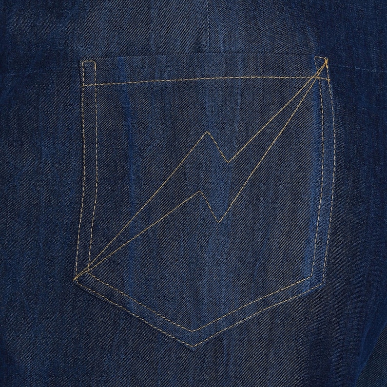 May include: Close-up of a dark blue denim pocket with a lightning bolt design stitched in a light tan thread. The pocket is rectangular with a curved bottom and is sewn onto the denim fabric with matching thread.