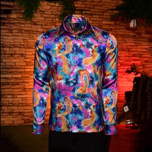 May include: A long-sleeve button-down shirt with a vibrant paisley pattern. The shirt features a colorful tie-dye background in shades of blue, purple, and pink, with gold and orange paisley designs. The shirt has a wide collar and buttoned cuffs.