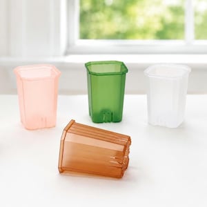 May include: Four square-shaped plastic plant pots in translucent colors. Three pots stand upright in light pink, green, and white, while one orange pot lies on its side in the foreground. Each pot features vertical ridges and drainage slots at the base.