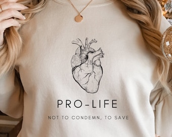 May include: Beige sweatshirt featuring a detailed black and white anatomical heart illustration. Below the graphic, the text reads "PRO-LIFE" above "NOT TO CONDEMN, TO SAVE". The sweatshirt has a crew neck and long sleeves.
