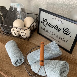 May include: A black wire basket with gray cloths and white laundry balls. A sign that reads "Pickering Laundry Co. Wash, Dry and Fold Self-Serve Open 24 Hours" is on a wooden surface with blue towels in a wire basket.
