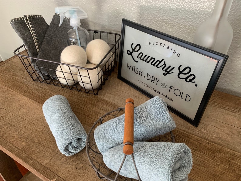 May include: A black wire basket with gray cloths and white laundry balls. A sign that reads "Pickering Laundry Co. Wash, Dry and Fold Self-Serve Open 24 Hours" is on a wooden surface with blue towels in a wire basket.