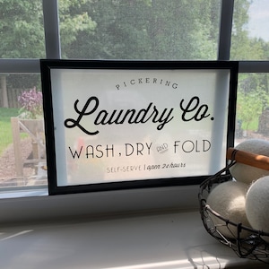 May include: A black framed sign with white text that reads "PICKERING Laundry Co. WASH, DRY AND FOLD SELF-SERVE | open 24 hours".