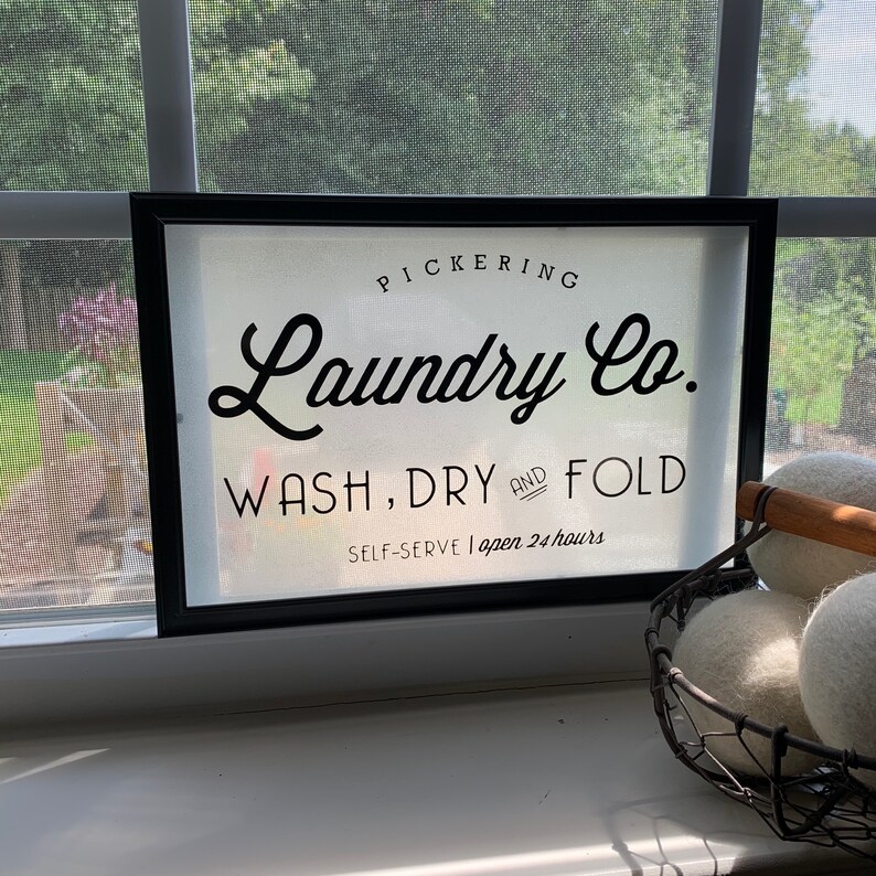 May include: A black framed sign with white text that reads "PICKERING Laundry Co. WASH, DRY AND FOLD SELF-SERVE | open 24 hours".