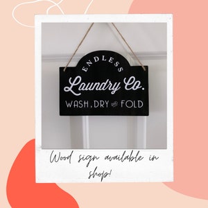 May include: Black wooden sign with white text that reads "Endless Laundry Co. Wash, Dry and Fold". The sign is hanging from a string and is in front of a white wall.