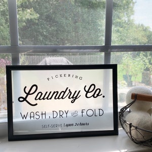 May include: A black framed sign with white lettering that reads "PICKERING Laundry Co. WASH, DRY AND FOLD SELF-SERVE | open 24 hours".