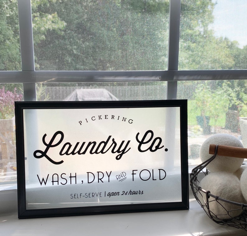 May include: A black framed sign with white lettering that reads "PICKERING Laundry Co. WASH, DRY AND FOLD SELF-SERVE | open 24 hours".