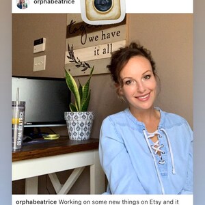 May include: A woman wearing a light blue shirt with white laces is working on new items for Etsy. The text "we have it all" is on a wooden sign behind her. A potted snake plant is on a table in front of her.