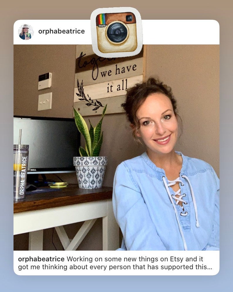 May include: A woman wearing a light blue shirt with white laces is working on new items for Etsy. The text "we have it all" is on a wooden sign behind her. A potted snake plant is on a table in front of her.