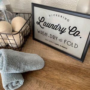 May include: A black framed sign with white background and black text that reads "PICKERING Laundry Co. WASH, DRY AND FOLD SELF-SERVE I open 24 hours". The sign is sitting on a wooden surface with a basket of white wool dryer balls and two rolled up gray towels.