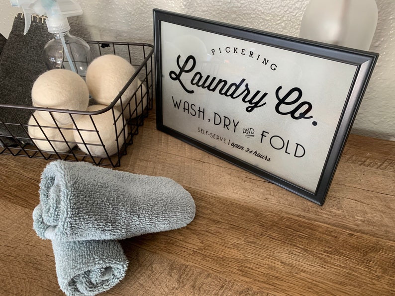 May include: A black framed sign with white background and black text that reads "PICKERING Laundry Co. WASH, DRY AND FOLD SELF-SERVE I open 24 hours". The sign is sitting on a wooden surface with a basket of white wool dryer balls and two rolled up gray towels.