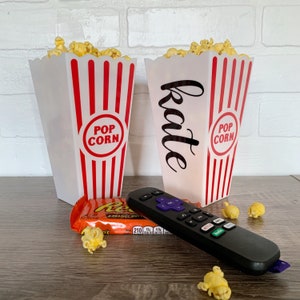 May include: Two red and white striped popcorn containers with the words "Pop Corn" on them. One container says "Katie" on it. A remote control and a bag of peanut butter cups are on a wooden surface.
