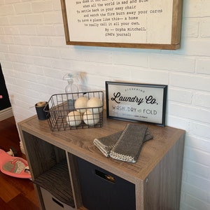 May include: A white brick wall with a wooden shelf holding a black metal basket with white pom poms, a clear spray bottle, and a black framed sign that reads "Pickering Laundry Co. Wash. Dry. Fold".