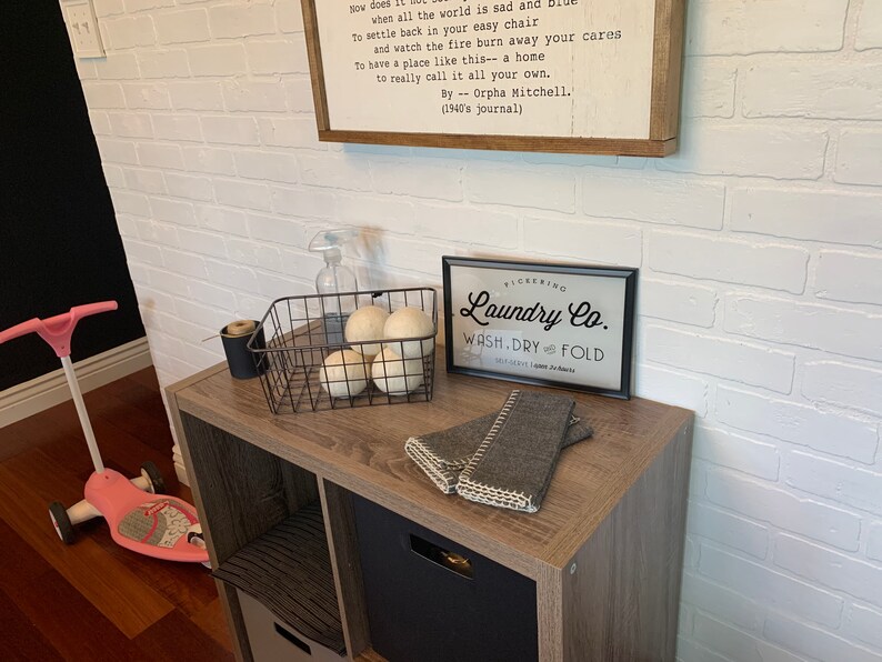 May include: A white brick wall with a wooden shelf holding a black metal basket with white pom poms, a clear spray bottle, and a black framed sign that reads "Pickering Laundry Co. Wash. Dry. Fold".