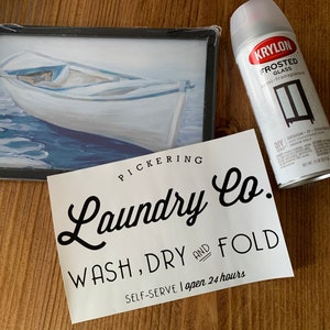 May include: A white sign with black text that reads "PICKERING Laundry Co. WASH, DRY AND FOLD SELF-SERVE I open 24 hours". A can of Krylon Frosted Glass spray paint is in the background.