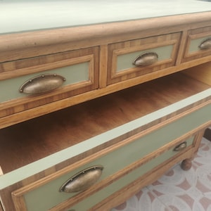 May include: A wooden dresser with four drawers and antique-style metal pulls. The dresser is painted a light green color and has a wood grain pattern on the drawers. The top drawer is open, revealing the wooden interior.