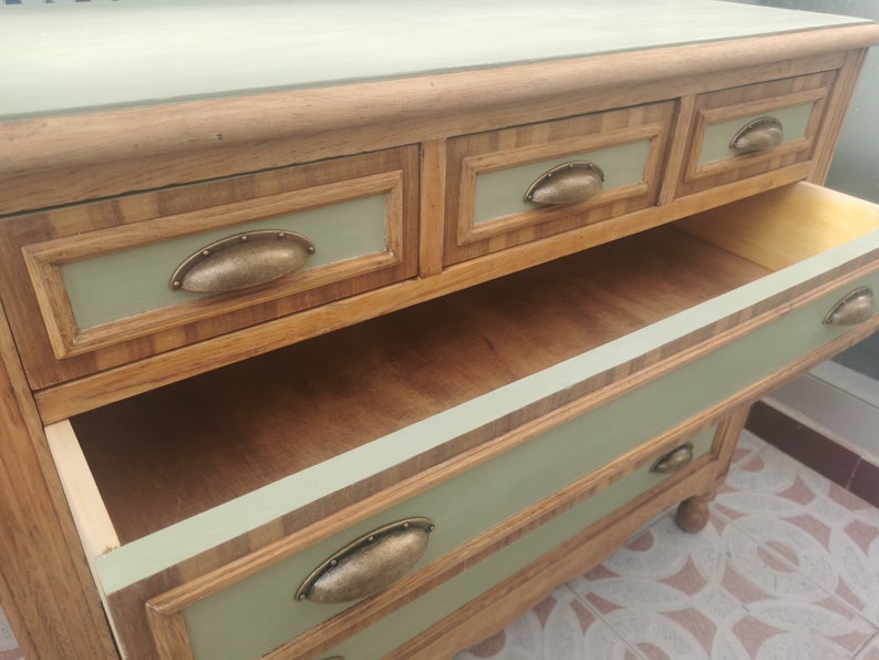 May include: A wooden dresser with four drawers and antique-style metal pulls. The dresser is painted a light green color and has a wood grain pattern on the drawers. The top drawer is open, revealing the wooden interior.
