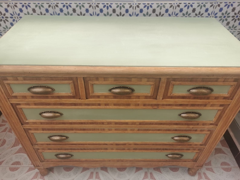 May include: A five-drawer dresser with a light green top and wooden drawers. The drawers have a light brown wood veneer with a woven pattern and brass oval-shaped handles.