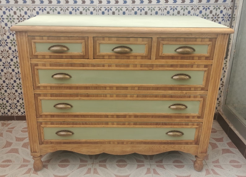 May include: A wooden dresser with six drawers, each with a brass handle. The dresser is painted a light green color and has a wood grain pattern. The dresser is sitting on a tiled floor.