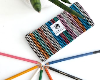 May include: A colorful striped pencil case with a zipper closure. The case is made of woven fabric and has a small logo that says "Finidi, empowering women". The case is surrounded by eight colored pencils.