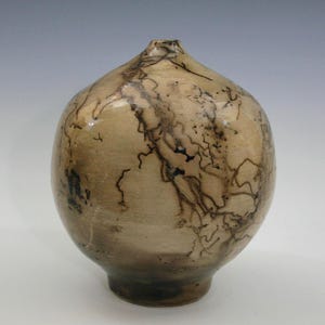 May include: A handcrafted ceramic vase with a rounded body and a narrow neck. The vase features a light brown glaze with dark, lightning-like patterns. The base is a darker shade of brown. A decorative piece for the home.