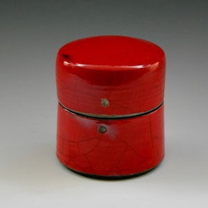 May include: A cylindrical, red ceramic container with a glossy finish. The container is composed of two stacked sections, with a lid on top. The surface has a crackled texture and small, circular accents.
