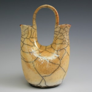 May include: A ceramic vase with a crackle glaze finish. The vase has two openings and a handle. The vase is a light cream color with brown and black crackle lines.