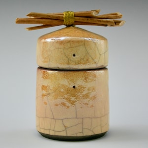 May include: A small, two-piece ceramic jar with a crackled glaze finish. The lid is topped with a bundle of twigs tied together with gold string.