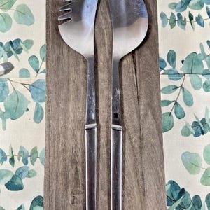 May include: A stainless steel salad serving set with a slotted spoon and a solid spoon, resting on a wooden serving board. The set is displayed on a patterned tablecloth with green and white foliage.