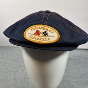 May include: A dark navy blue flat cap featuring an oval embroidered patch on the front. The patch has a yellow border and contains the text Chevrolet Corvette in brown, along with a red flag and a black and white checkered flag graphic.