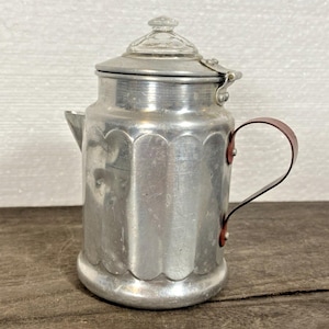 May include: A vintage aluminum coffee pot with a clear glass knob on the lid. The pot has a scalloped design around the base and a brown handle. The pot is approximately 6 inches tall.