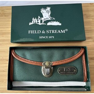 May include: A dark green rectangular wallet with brown trim and a metal clasp closure. The wallet features a metal plaque with the text F&S FIELD & STREAM. It is presented in a dark green box with a white eagle logo and the text FIELD & STREAM SINCE 1871.